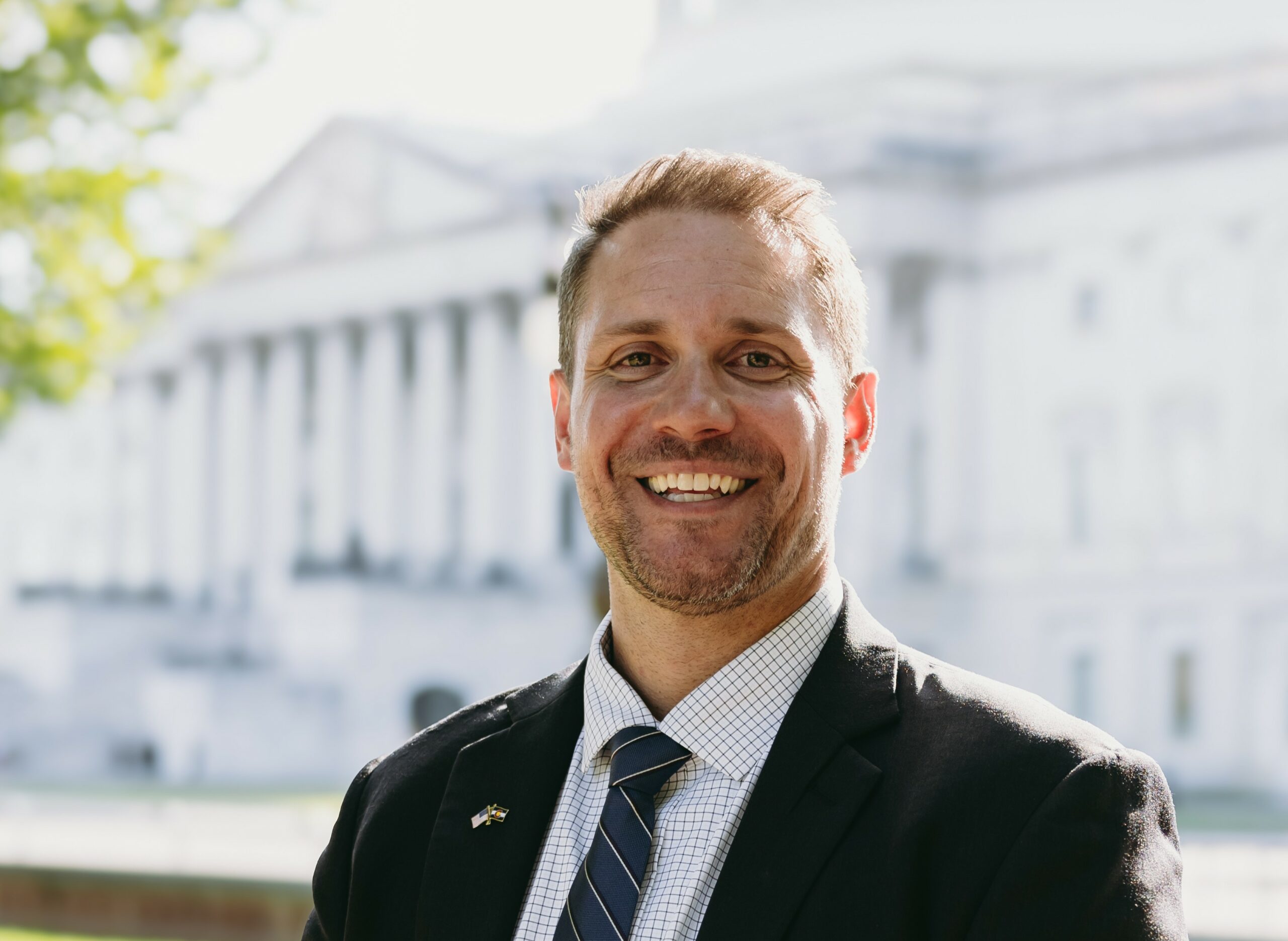 "A smiling man in a suit and tie standing outdoors in front of a government-style building with white columns