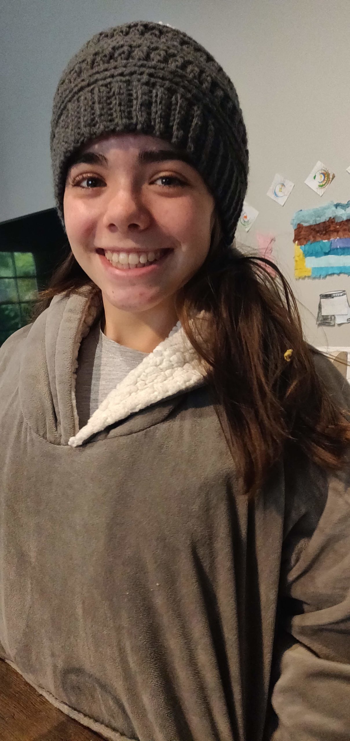 Female with brown hair wearing a grey knit hat and brown top smiling at the camera